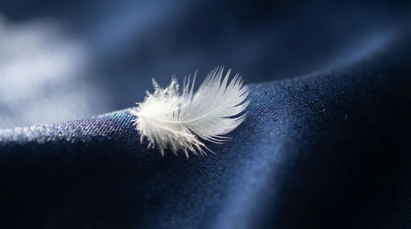Close-up macro photo of a tiny white feather on a rich navy blue silk background.
