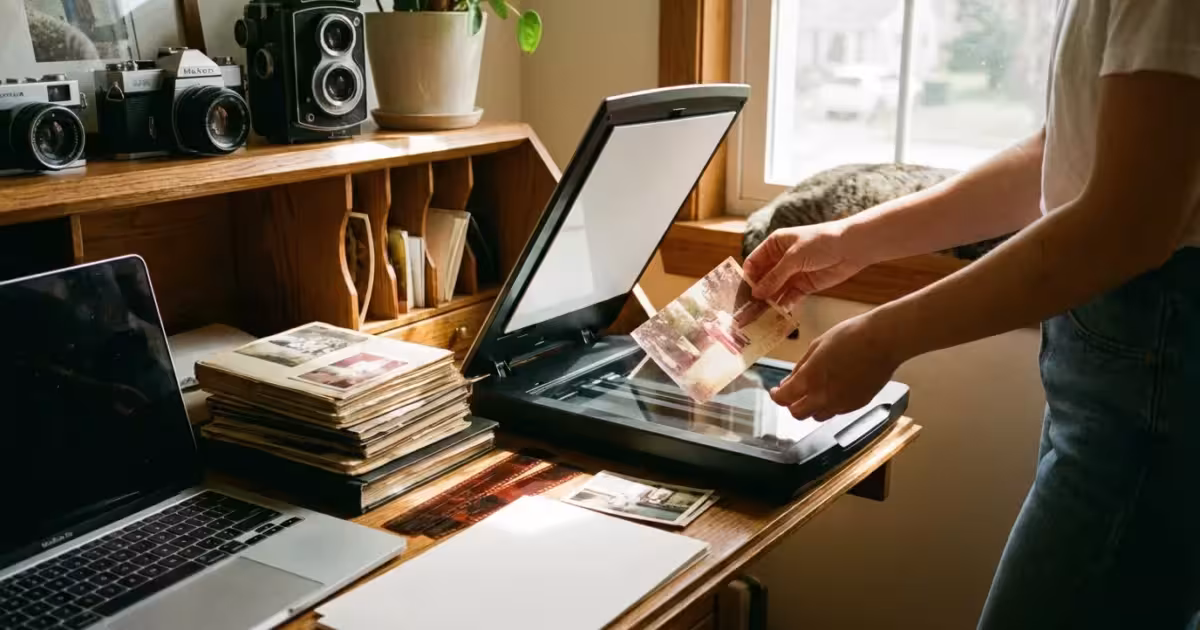 A person scanning a vintage family photograph on a modern flatbed scanner in a sunlit home office.