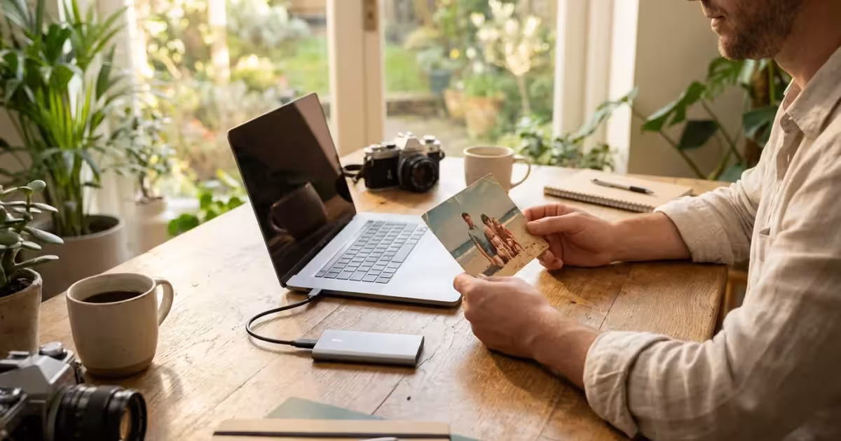 A photographer holding a physical photo next to a modern laptop and external SSD on a wooden desk.