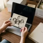 A person holding a vintage family photo over a laptop, symbolizing the transition from physical to digital archives.