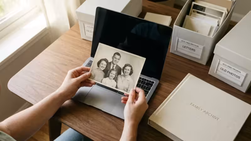 A person holding a vintage family photo over a laptop, symbolizing the transition from physical to digital archives.