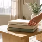 A stack of high-quality linen photo books on a wooden table in a bright room.