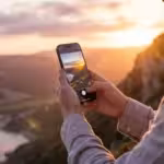 A person using a smartphone to photograph a golden hour landscape in a high-end editorial style.